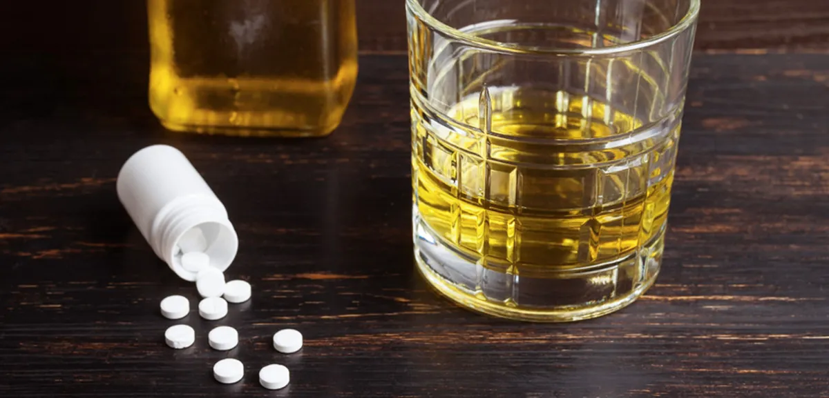 A glass and a bottle of liquor next to spilled white pills on a dark wood table